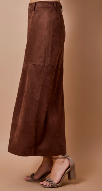 A brown wide-leg faux-suede skirt shown on a model from the waist down, paired with taupe block-heel sandals against a solid beige studio background.