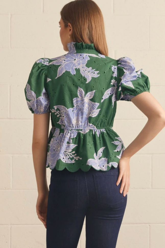A woman shown from the back wearing a green eyelet blouse with a lavender floral print and dark blue jeans, standing against a light beige brick wall.
