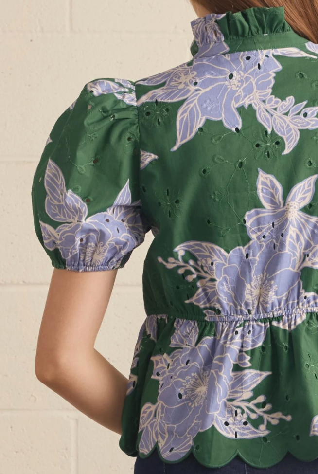 Close-up of a woman wearing a green eyelet blouse with lavender floral print, featuring puff sleeves, a ruffled high neck, and a scalloped peplum hem.
