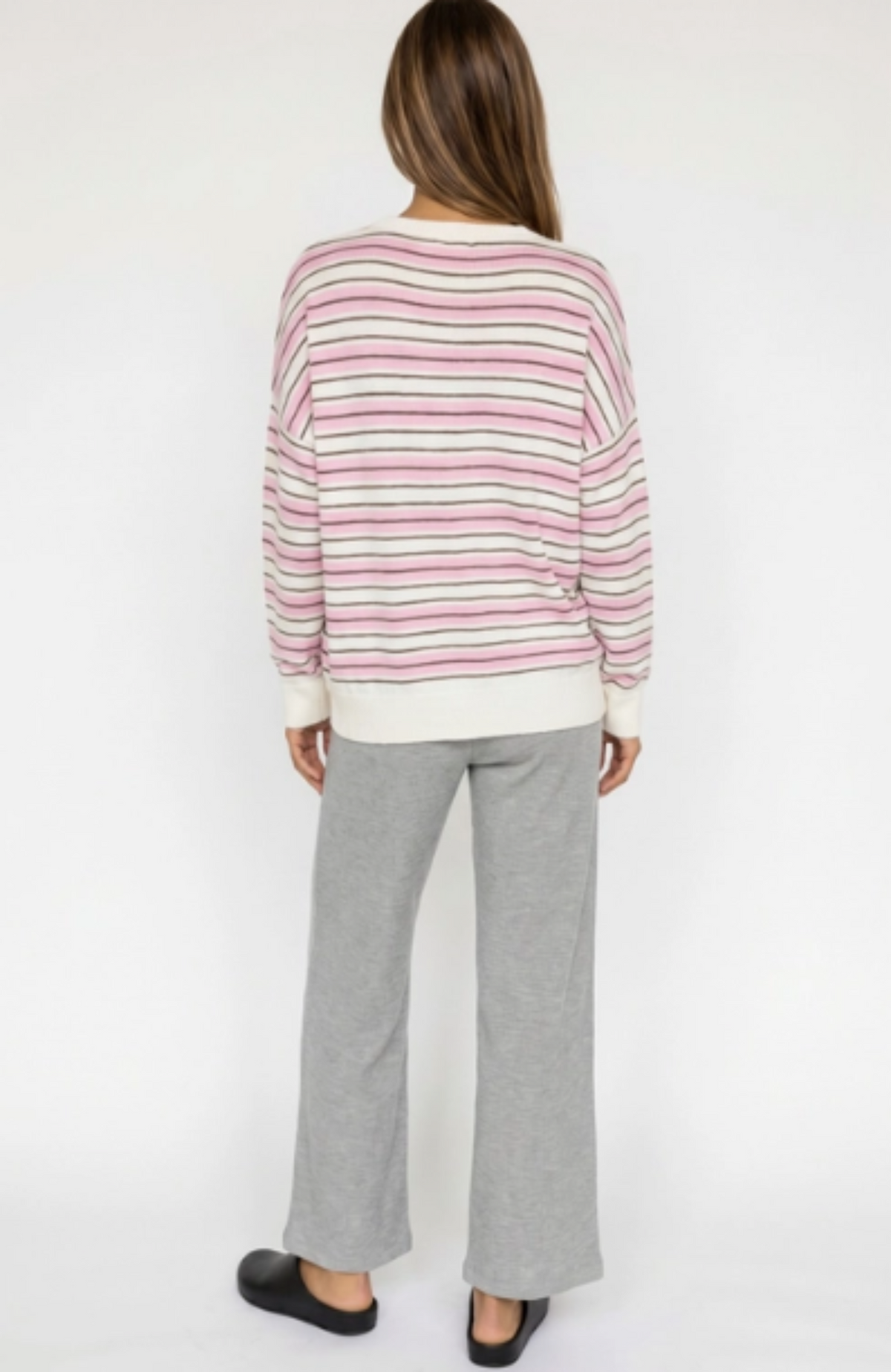 A woman shown from the back wearing a pink and white striped long-sleeve knit top with gray wide-leg sweatpants and black slip-on shoes, against a plain white studio background.