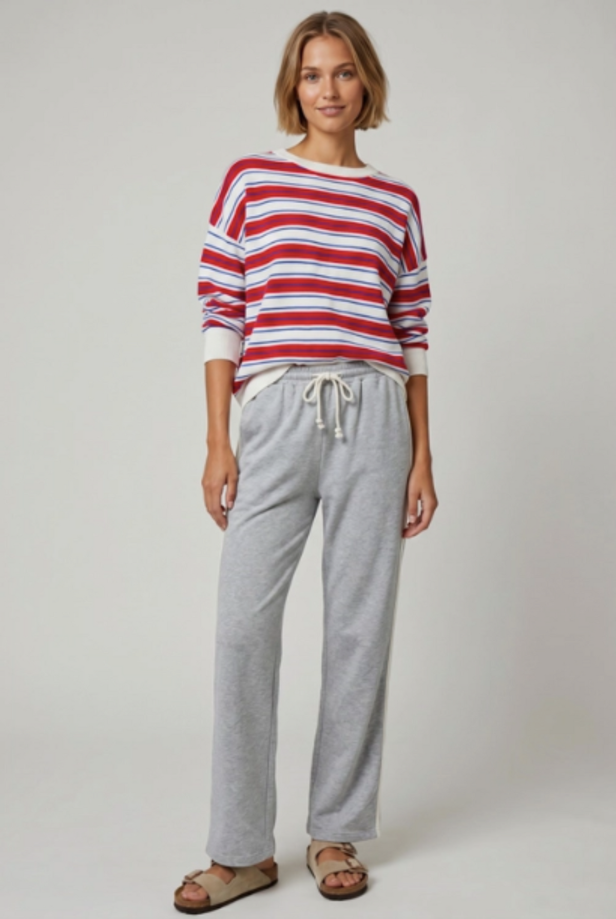 A woman modeling a red, white, and blue striped long-sleeve knit top paired with light gray drawstring sweatpants and brown sandals, photographed against a plain light gray studio background.