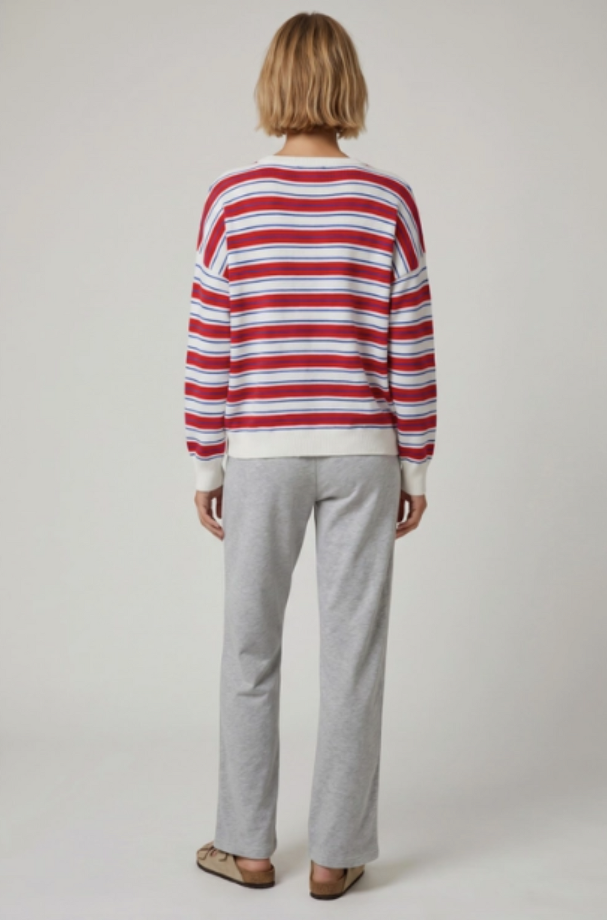 A model viewed from behind wearing a red, white, and light blue horizontally striped long-sleeve crewneck sweater and light gray sweatpants against a neutral light gray background.