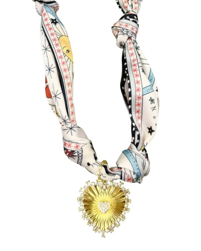 A patterned silk scarf styled as a necklace with a gold heart-shaped pendant featuring a clear crystal center, set against a plain white background.
