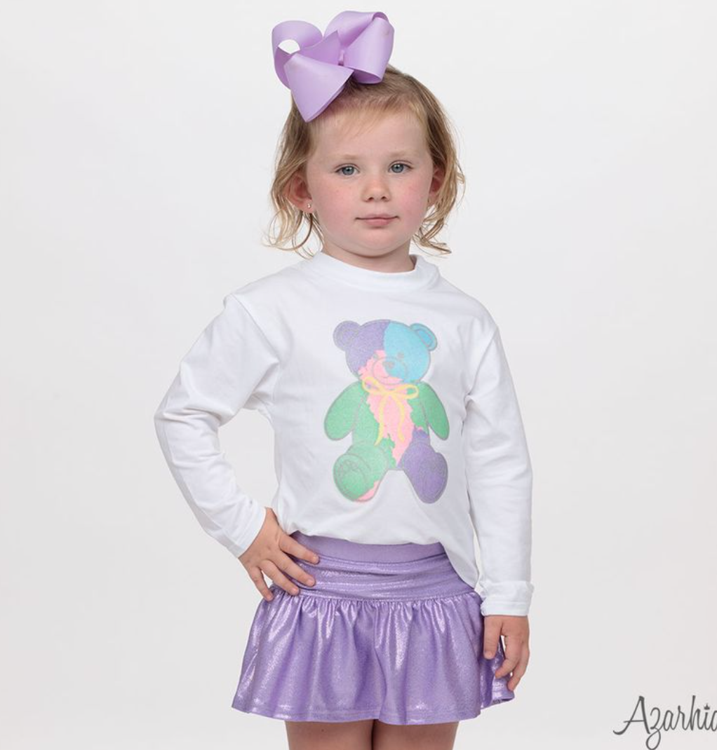 A young girl with a large lavender bow in her hair stands against a plain white studio background, wearing a white long-sleeve top with a colorful teddy bear graphic and a shiny lavender skirt.
