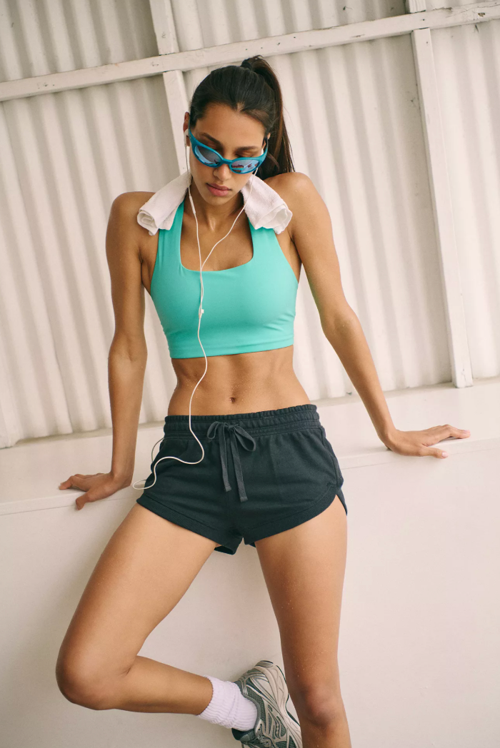 A woman in athletic wear posing against a white corrugated metal wall, wearing a mint green sports bra, black athletic shorts, white socks, gray running shoes, and blue-tinted sunglasses with a white towel around her neck.