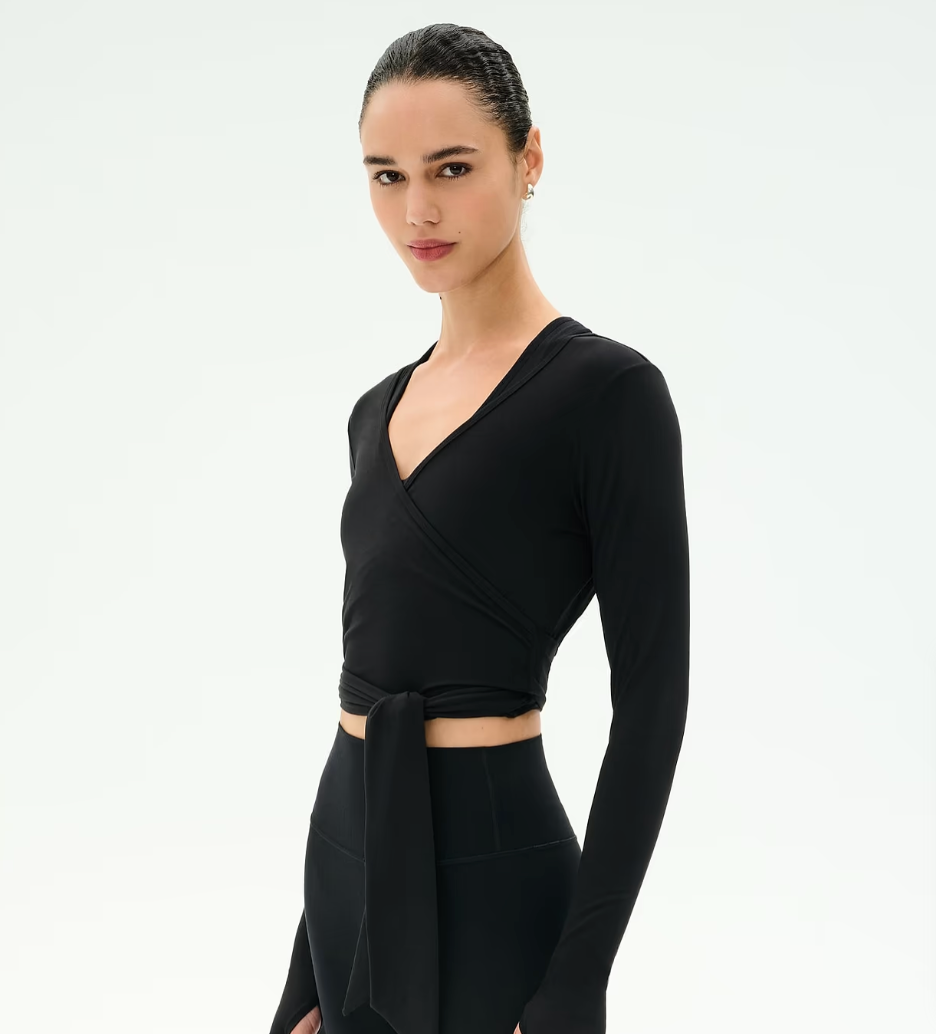 A woman modeling a black long-sleeve wrap-style crop top with a side tie detail, paired with black high-waisted leggings, against a plain white studio background.