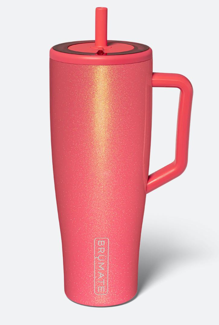 A coral pink Primalate insulated travel tumbler with a matching handle, lid, and straw, featuring a subtle gold glitter finish and the Primalate logo near the bottom, set against a light gray background.