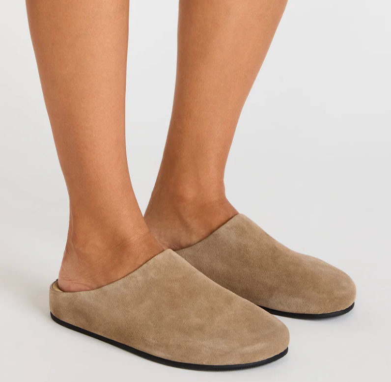 A pair of taupe suede slip-on mule slippers worn on a model's feet, shown against a plain light gray studio background.
