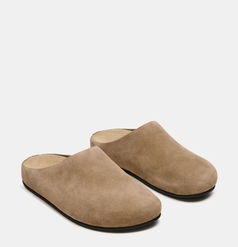 A pair of taupe suede mule slip-on shoes with black soles, displayed on a clean light gray background.