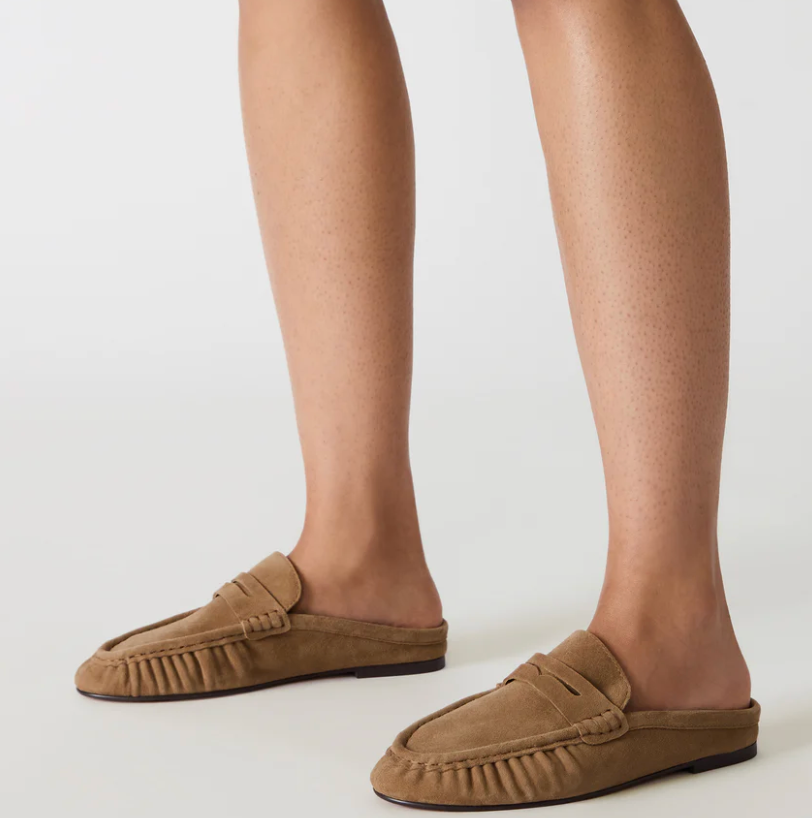 A pair of taupe-brown suede mule loafers shown on a model's feet against a clean, light gray studio background.
