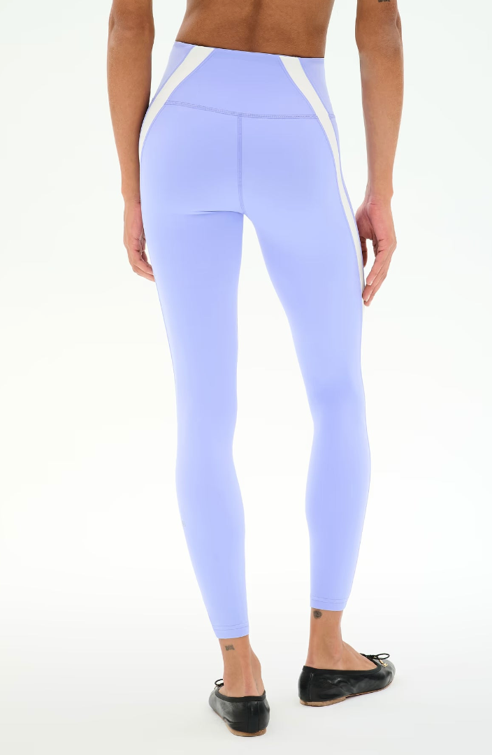 A model from the back wearing light purple high-waisted leggings with white side panels, paired with black flats, against a plain white background.