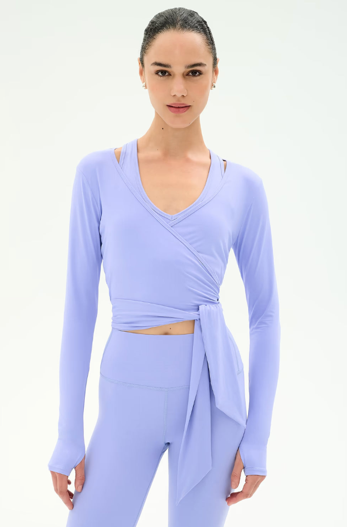 A woman modeling a light lavender long-sleeve wrap-style top with a side tie and matching high-waisted leggings against a plain white studio background.
