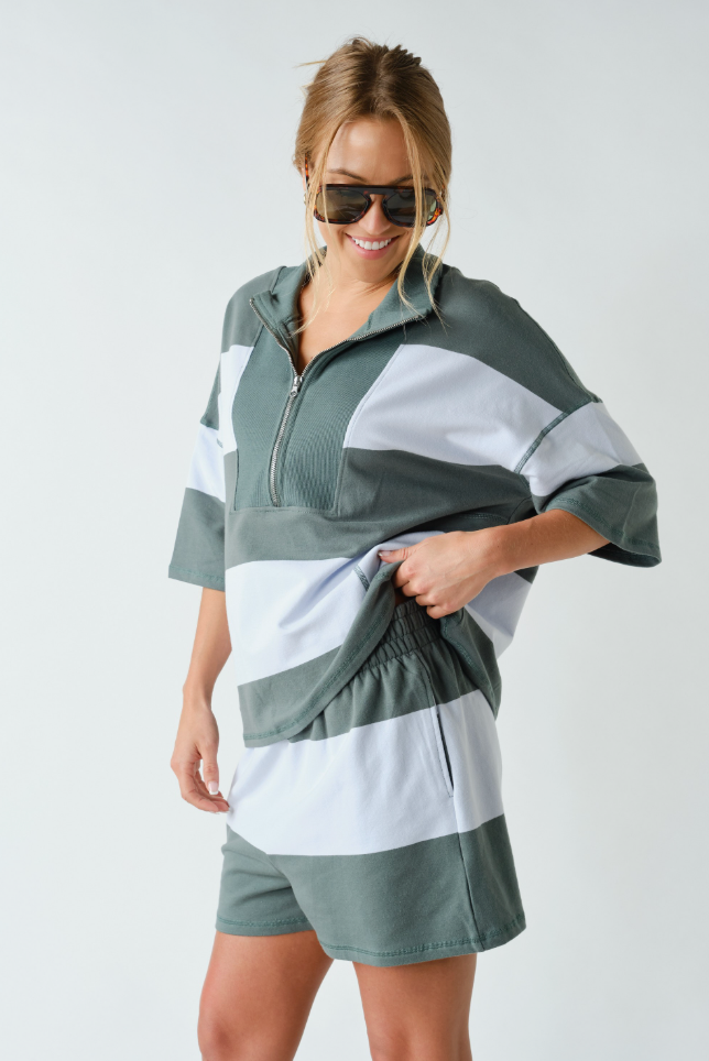 A woman modeling a matching two-piece lounge set with a half-zip pullover top and shorts in a color-blocked green and white design, set against a plain light gray background.