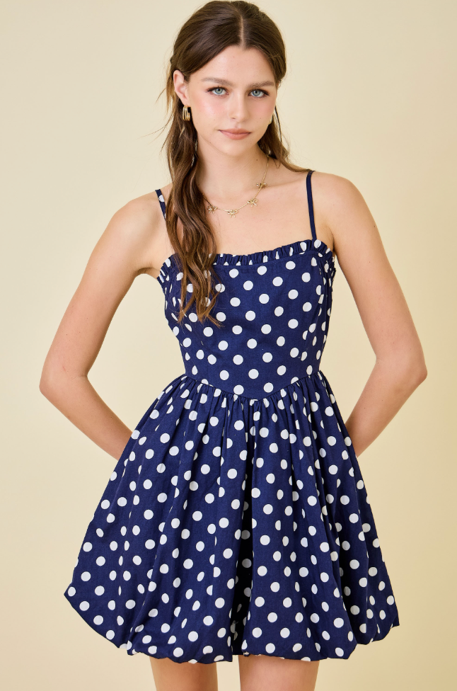 A young woman modeling a navy blue and white polka dot spaghetti-strap mini dress against a solid beige background.