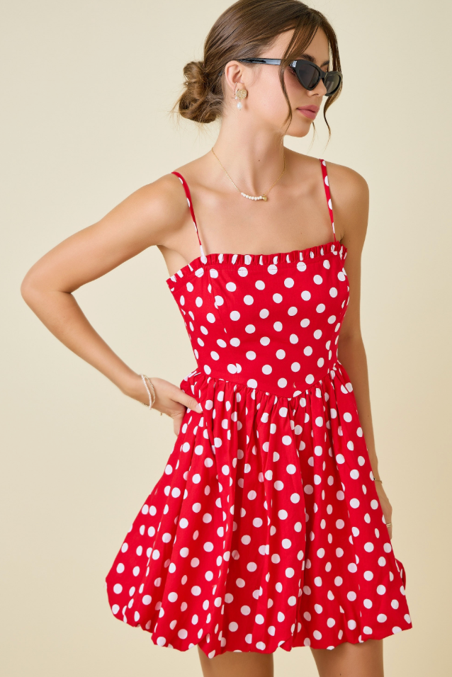 A woman modeling a red spaghetti-strap mini dress with white polka dots, styled with sunglasses, a necklace, and bracelets against a solid beige background.