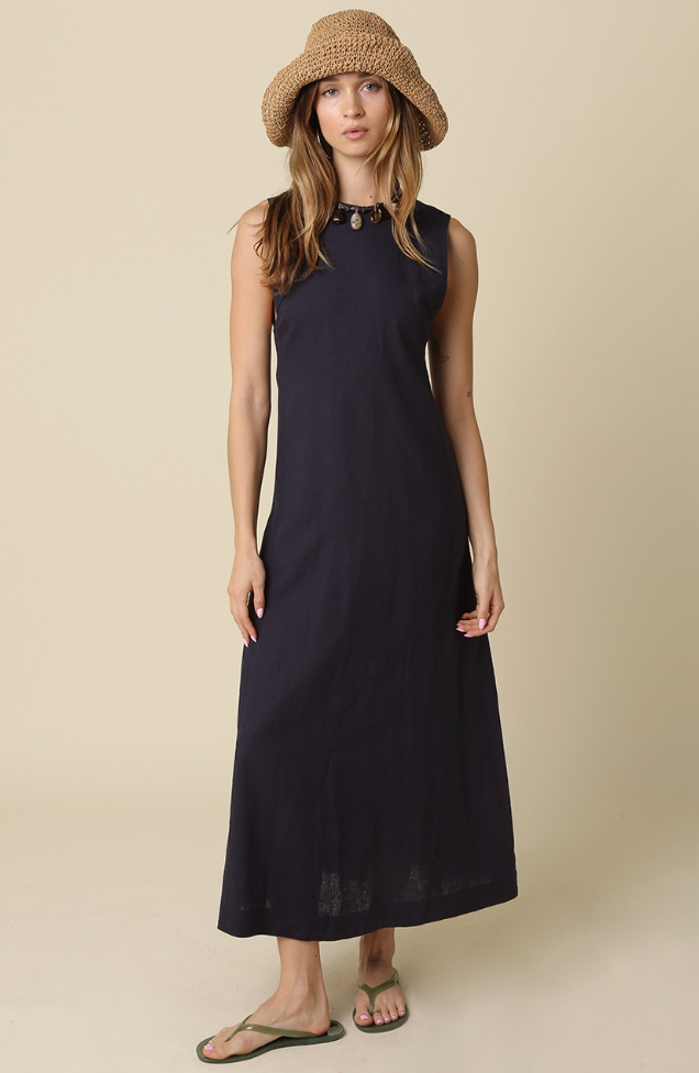 A woman modeling a sleeveless navy maxi dress with a high neckline and a woven straw hat, standing against a solid beige background.