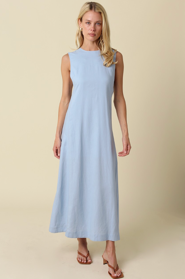 A woman modeling a sleeveless light blue maxi dress with a high round neckline, photographed against a solid beige studio background.