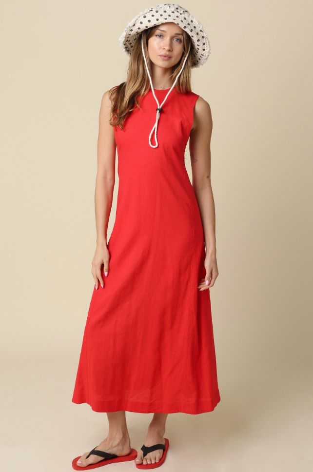A woman modeling a sleeveless red midi dress with a white polka-dot sun hat and red flip-flops against a solid beige studio background.