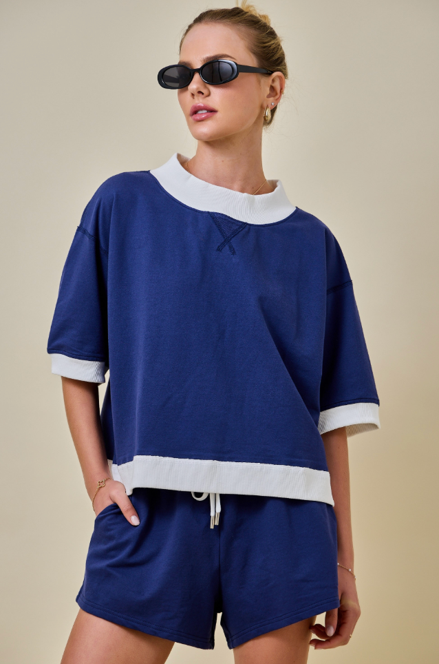 A woman modeling a navy blue two-piece lounge set with white contrast trim, featuring a short-sleeve top with a wide collar and hemband, and matching shorts, against a solid beige background.
