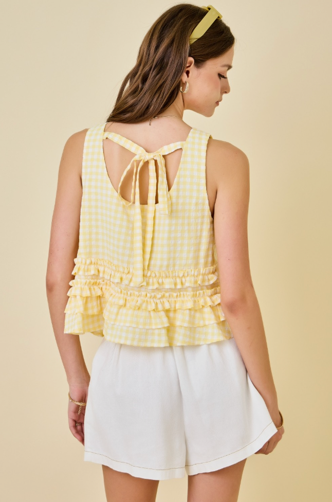 A woman shown from the back wearing a pale yellow and white gingham sleeveless top with a tie-back neckline and ruffled hem, paired with white shorts against a warm beige studio background.