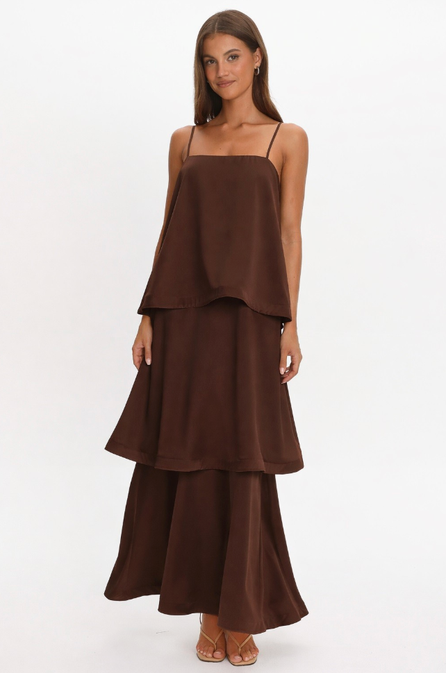 A woman modeling a dark brown two-piece set consisting of a spaghetti-strap camisole top with a layered ruffle hem and matching tiered maxi skirt, photographed against a seamless white studio background.