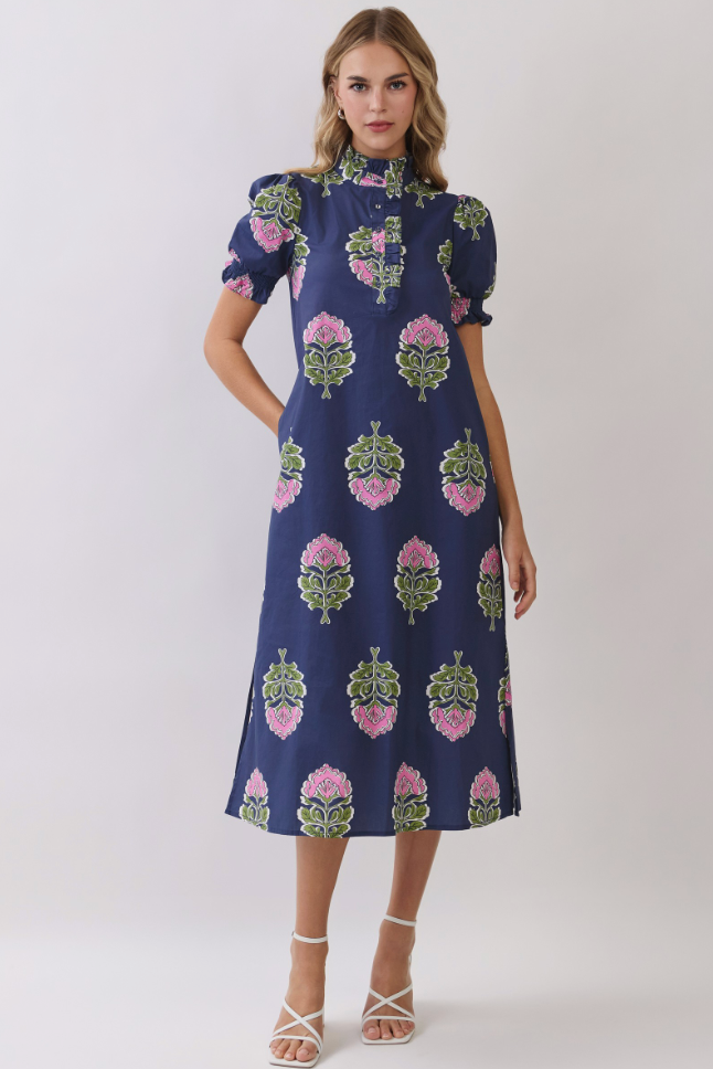A woman modeling a navy blue midi dress with puff sleeves, a high mock neck, and a pink and green floral block print pattern, paired with white strappy high-heeled sandals against a light gray studio background.