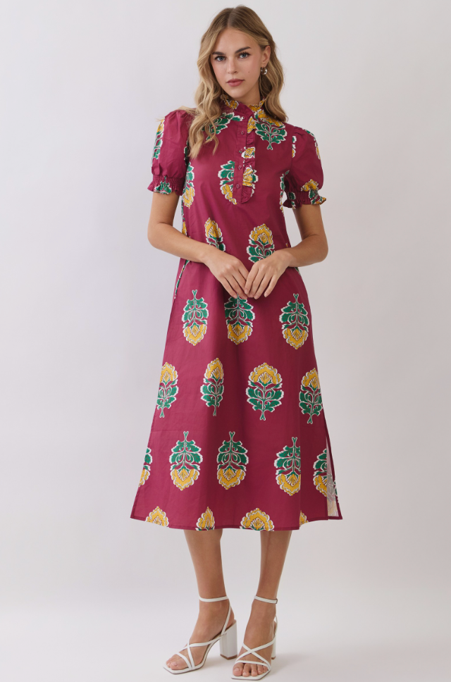 A woman modeling a short-sleeved midi dress with puff sleeves and an allover botanical print in green, yellow, and white on a deep burgundy base, set against a plain light gray studio background.