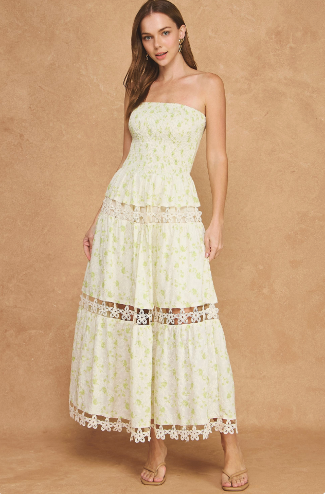 A woman modeling a strapless tiered maxi dress with a light cream and pale yellow floral print, lace trim, and a ruffled hem, set against a textured tan studio backdrop.