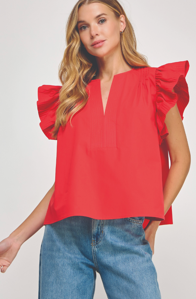 A woman wearing a bright red short-sleeve blouse with dramatic ruffled shoulders and a split neckline, paired with blue jeans, against a light gray studio background.