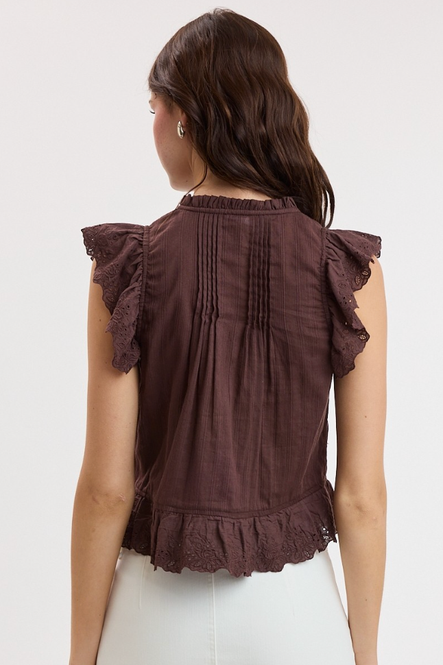 A woman viewed from the back wearing a dark brown sleeveless blouse with ruffled cap sleeves, vertical pleats, and a ruffled hem, paired with white pants against a plain light background.
