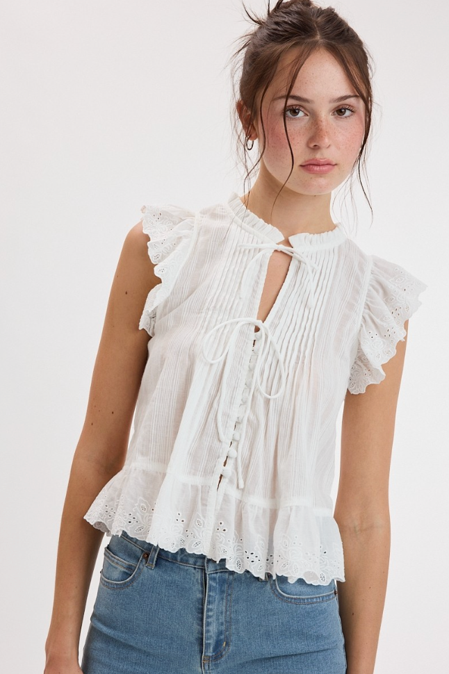 A woman wearing a white sleeveless blouse with ruffled shoulders, a keyhole tie neckline, and eyelet lace details, paired with blue jeans against a plain light background.