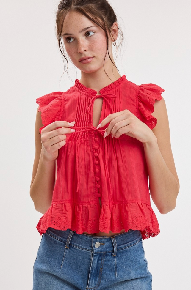 A woman wearing a sleeveless red ruffled blouse with a keyhole cutout and drawstring tie at the neckline, paired with blue jeans, against a plain light background.