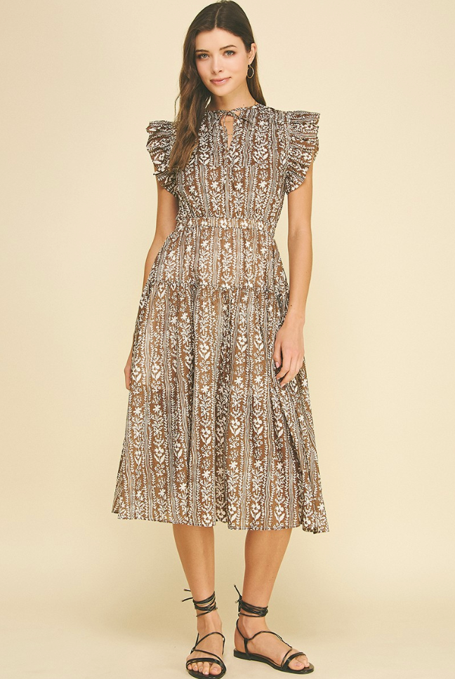 A woman modeling a midi-length dress with a beige and brown bohemian pattern, featuring ruffled cap sleeves and a high neckline, paired with black strappy sandals against a solid beige background.