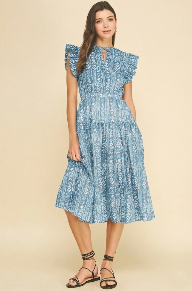 A woman modeling a light blue tiered midi dress with a white floral print, ruffled cap sleeves, and a keyhole neckline, paired with black strappy sandals against a solid beige background.