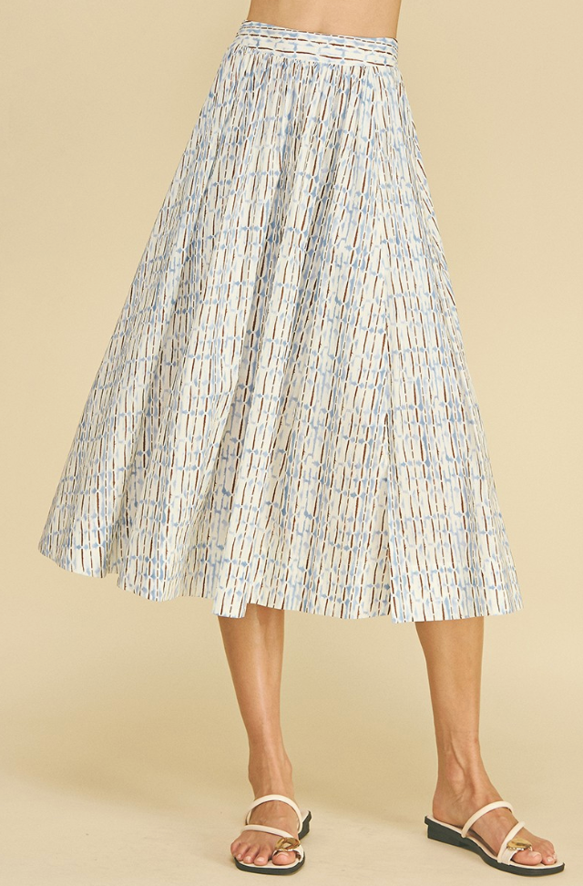 A pleated midi skirt with a white base and a light blue geometric pattern, shown on a model wearing white strappy sandals against a solid beige background.