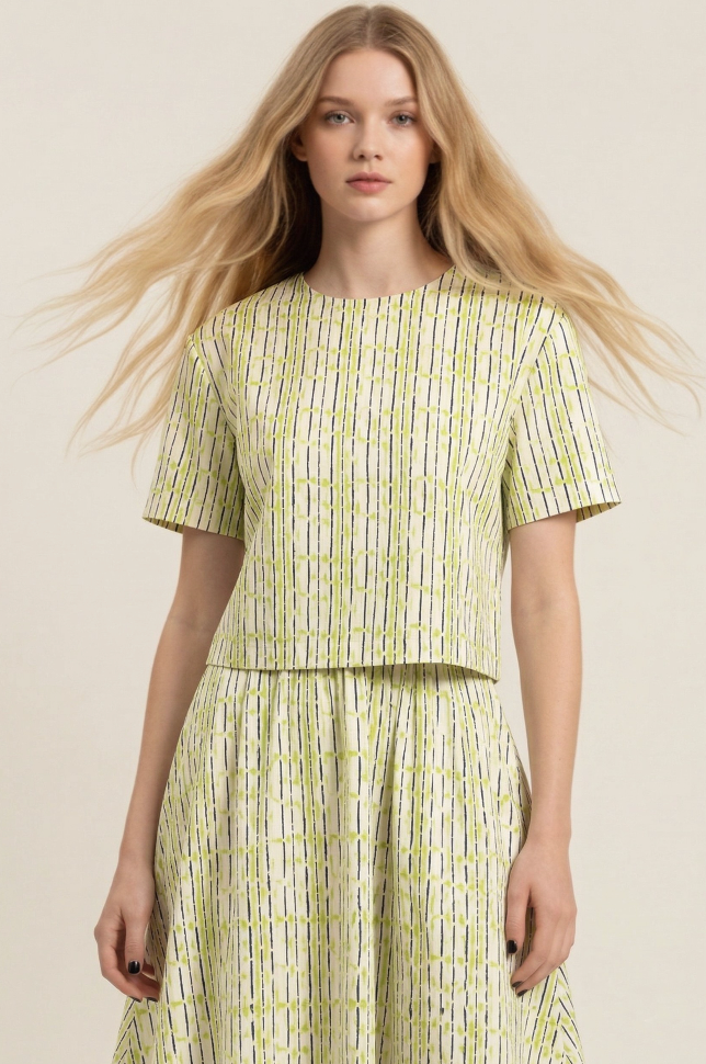 A woman modeling a matching short-sleeve crop top and pleated skirt set in a light cream fabric with a vertical lime green and black stripe pattern, set against a plain beige background.