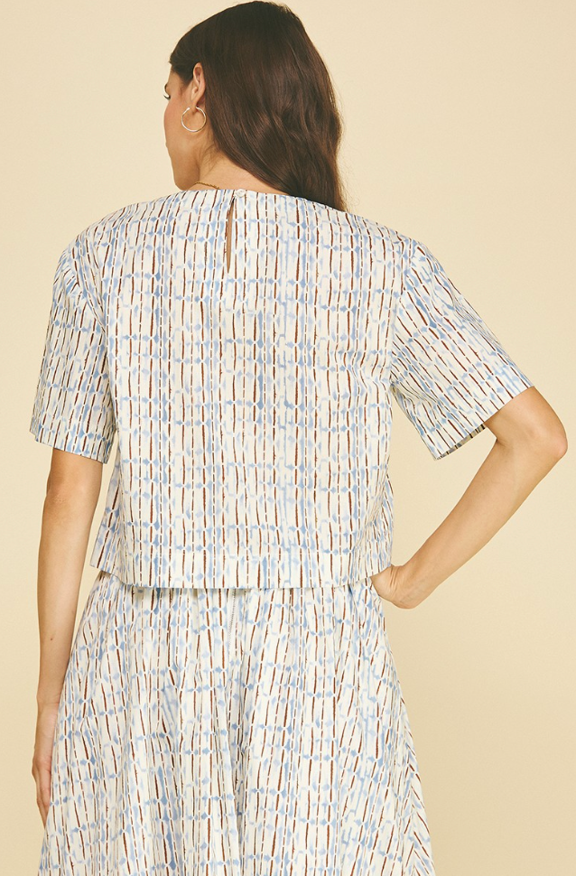 A woman shown from the back wearing a short-sleeve cropped top and matching skirt set with a vertical striped pattern in light blue, brown, and beige on an off-white base, against a solid beige background.