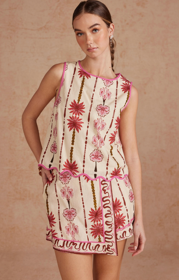 A sleeveless cream-colored mini dress with pink and brown botanical patterns, featuring pink piping and a ruffled hem, modeled against a textured beige background.