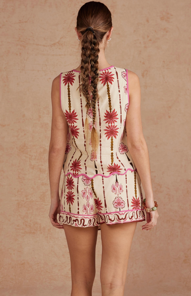A sleeveless cream-colored mini dress with pink and brown botanical patterns, featuring pink piping and a ruffled hem, modeled against a textured beige background.