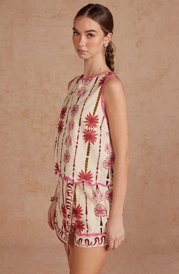A woman modeling a sleeveless cream-colored mini dress with pink and brown bohemian floral patterns, standing against a textured beige backdrop.
