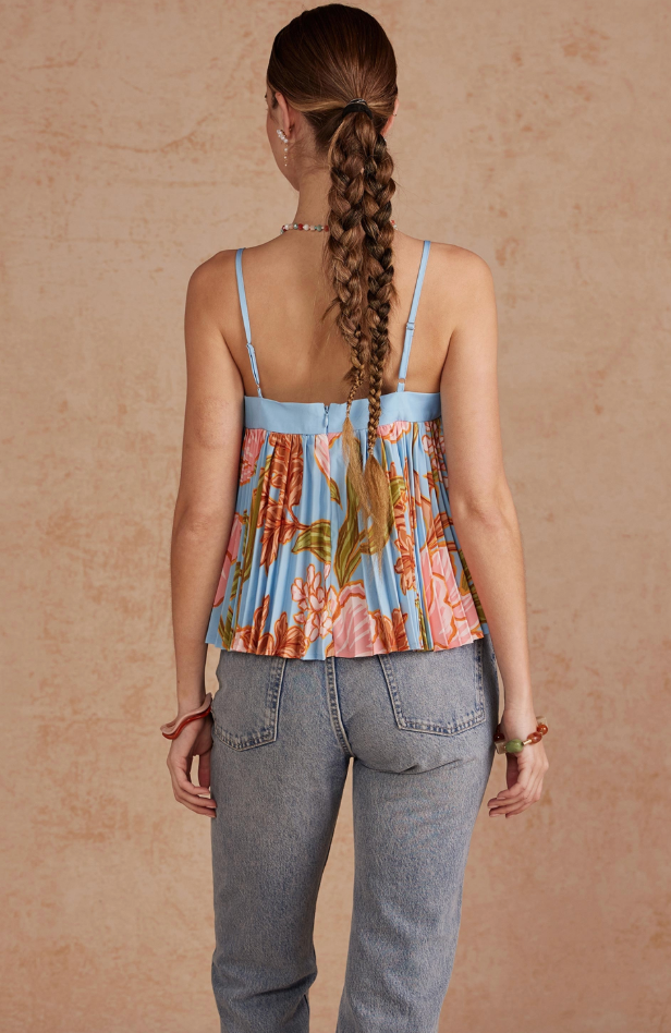 A woman viewed from behind wearing a light blue floral camisole top with gray jeans, standing against a textured beige studio backdrop.