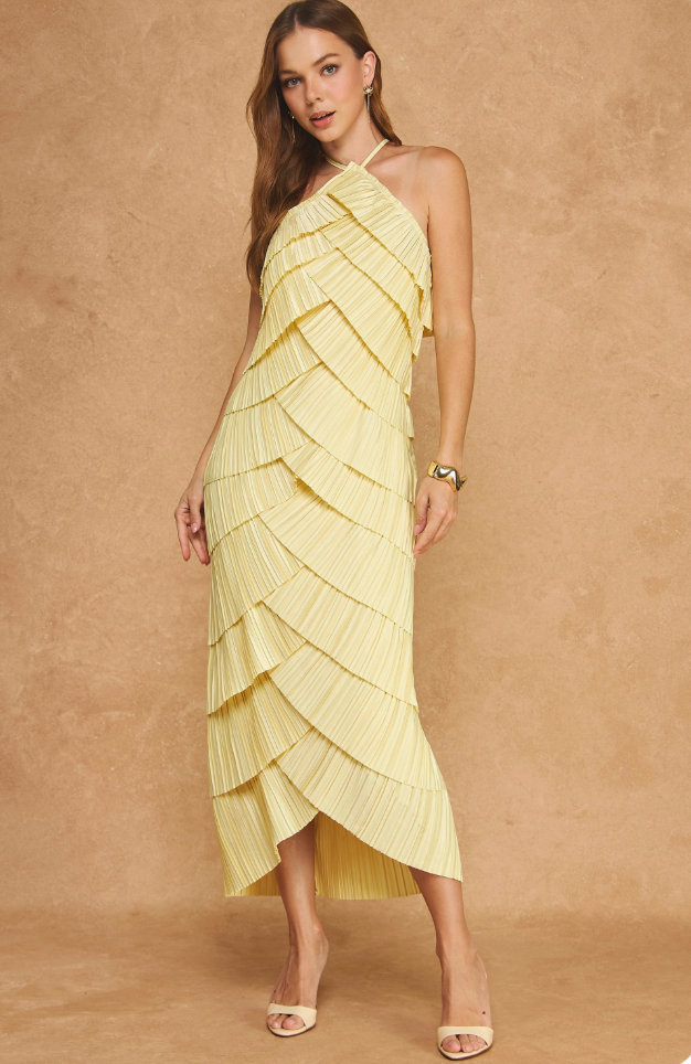 A woman models a pale yellow pleated halter midi dress with a ruffled, tiered overlay design against a textured beige studio backdrop.