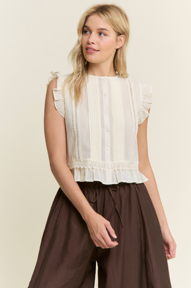 A woman wearing a cream sleeveless blouse with ruffle trim and button details, paired with dark brown wide-leg pants, against a plain beige background.