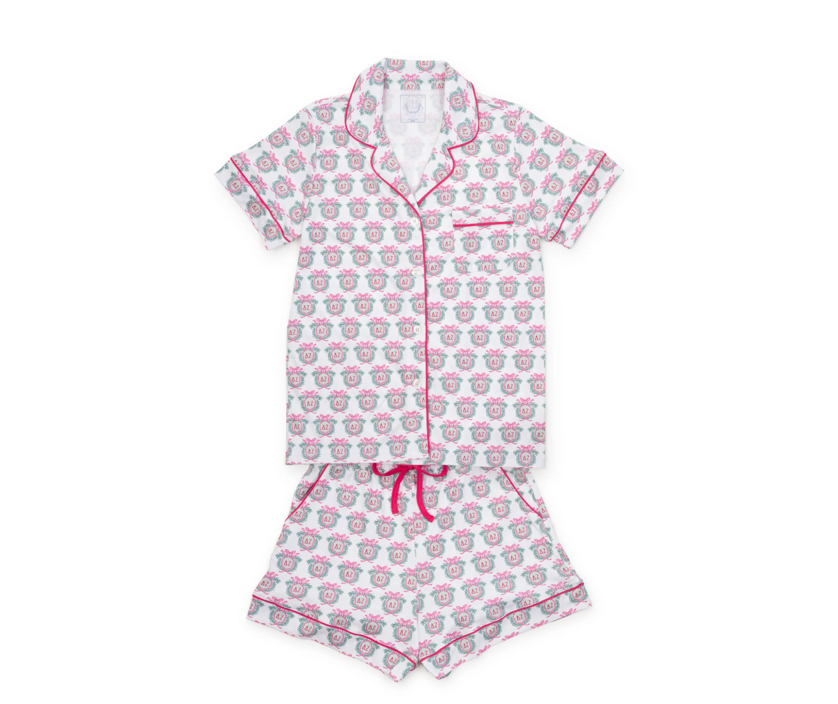 A two-piece girls' pajama set featuring a short-sleeve button-up top and matching shorts. The pajamas are white with a repeating pattern of pink cartoon sheep faces and are finished with pink piping and a pink drawstring waist.