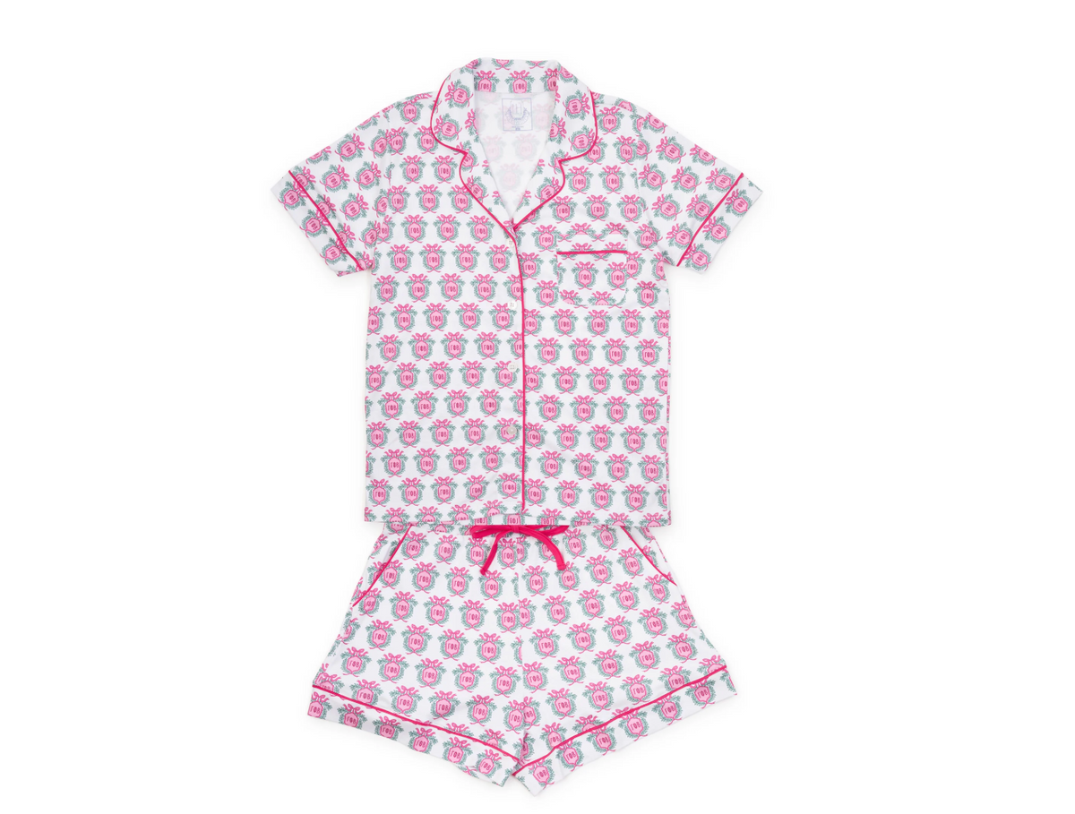 A two-piece girls' pajama set featuring a short-sleeve button-up top and matching shorts, both printed with a pink and gray cartoon face pattern and finished with pink piping and a pink drawstring.