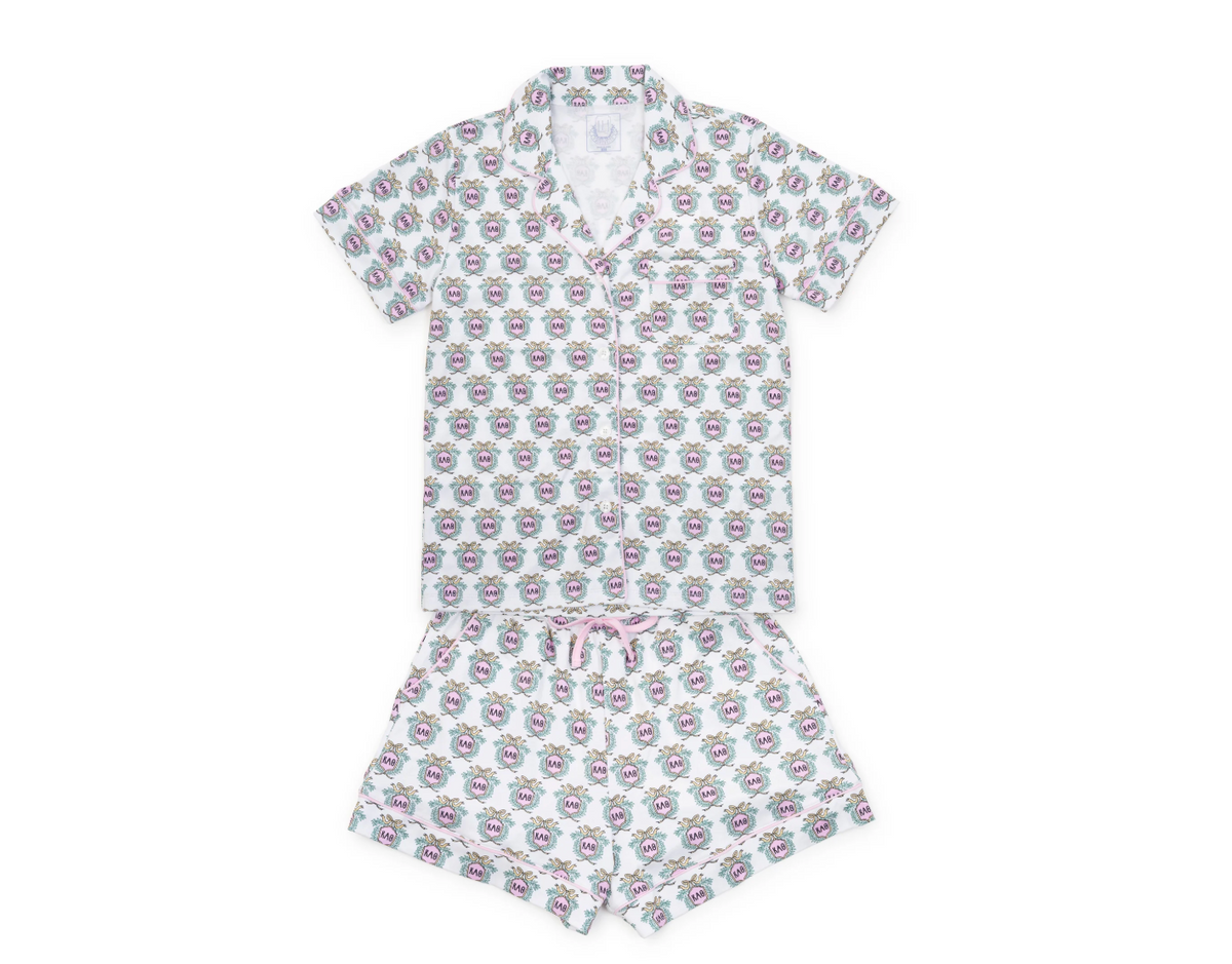 A two-piece short-sleeve pajama set laid flat on a white background, featuring a matching top and shorts in white with a repeating pink and purple cartoon face pattern and a small pink bow on the waist.