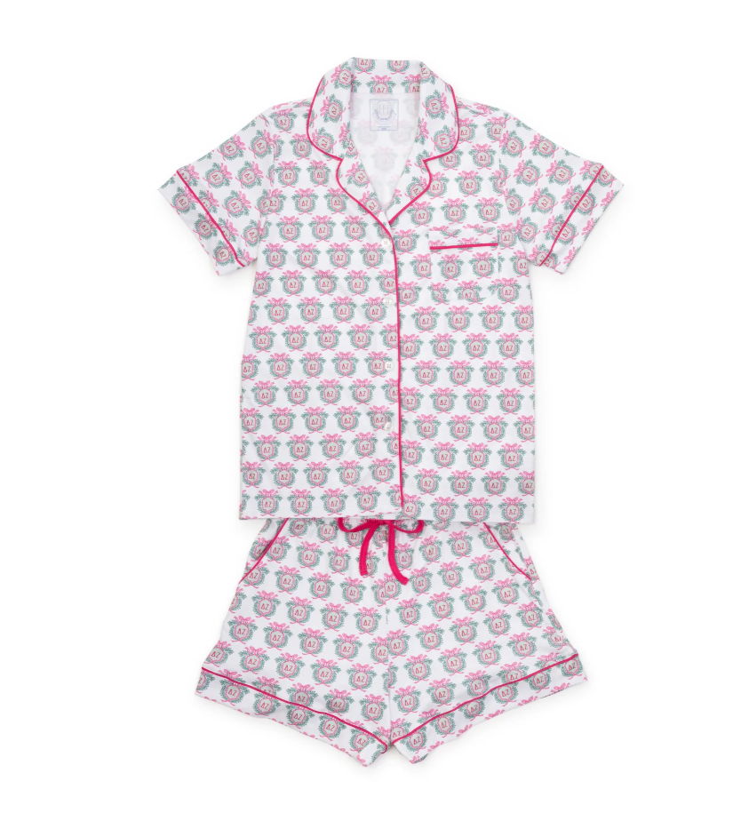 A two-piece baby pajama set featuring a short-sleeve button-up top and matching shorts. The pajamas are white with a repeating pattern of pink and gray cartoon sheep, finished with bright pink piping and a pink drawstring.