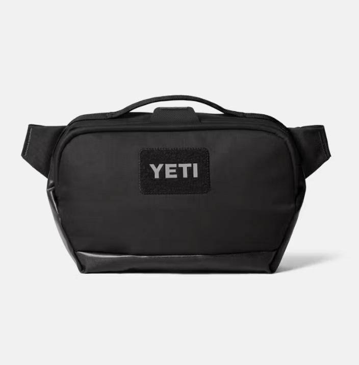 A black YETI shoulder bag with a top handle and a black rectangular patch on the front featuring the white YETI logo, set against a light gray background.