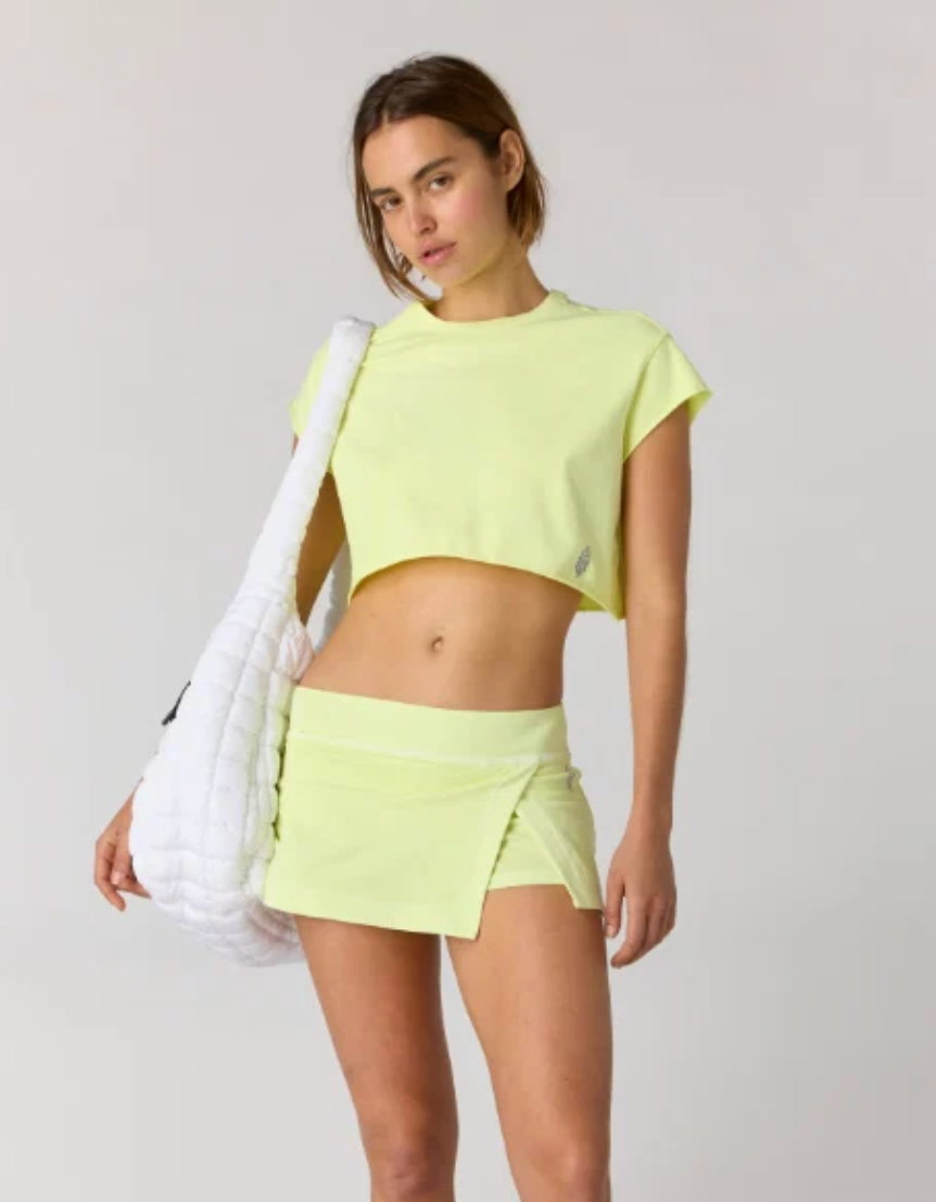 A model wearing a neon yellow-green athletic crop top and matching wrap-style mini skirt, carrying a large white quilted shoulder bag against a light gray studio background.