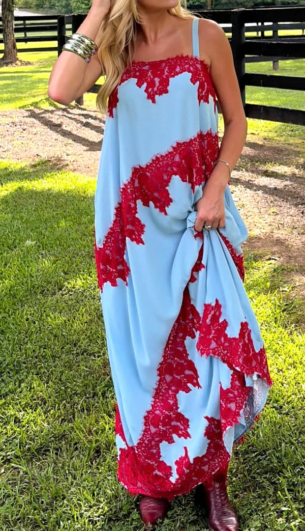A woman wearing a light blue maxi dress with red lace applique, standing outdoors on a grassy lawn with a wooden fence in the background.